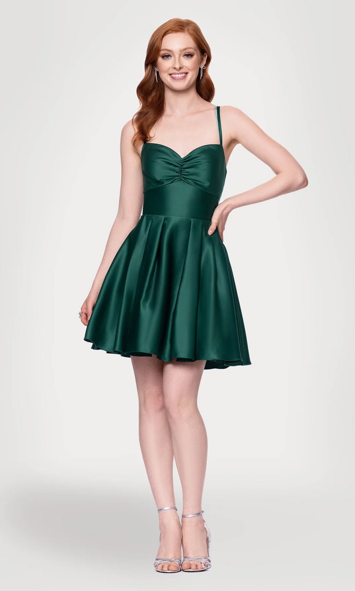 A woman with long red hair wears the Hunter Green Mini Hoco Dress: Blondie Nites 4927BN, a short green dress with thin straps, paired with silver high-heeled sandals, standing against a plain light background.